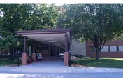 Covered entrance canopy with brick columns and benches leading to a single-story building surrounded by trees and lawn.