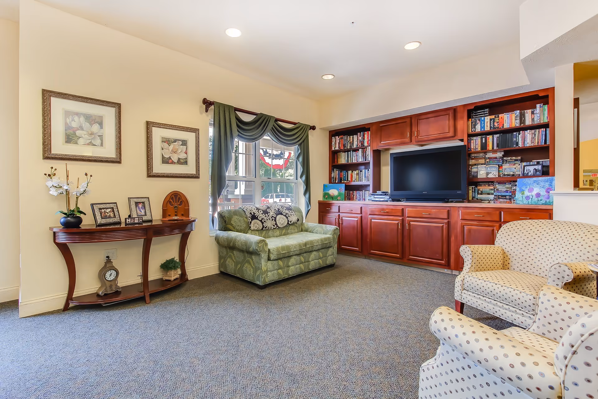 A cozy living room area in an assisted living facility featuring a green patterned loveseat, two beige armchairs with polka dots, a wooden console table with decorative items and framed pictures, a window with green drapes, and a built-in wooden bookshelf and cabinet unit with a flat-screen TV and numerous books and games.