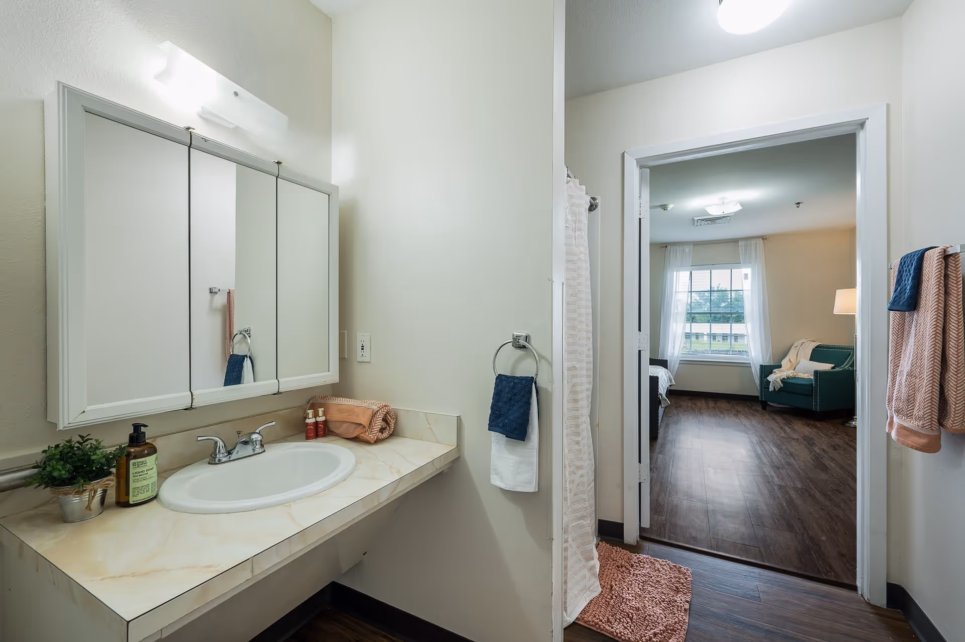 A bathroom with a sink, countertop, and mirror cabinet above. There are towels hanging on towel rings and a shower curtain partially visible. Through the open door, a bedroom with a window, green armchair, and lamp is visible.