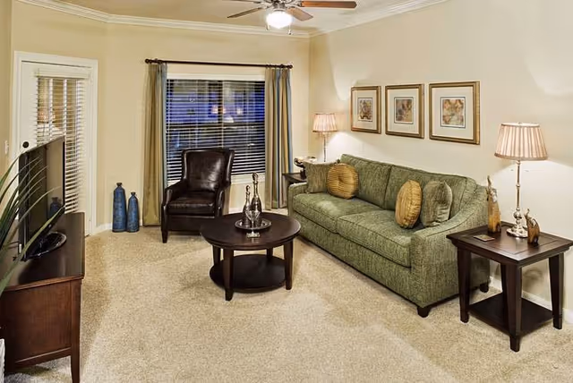 A cozy living room with a green sofa adorned with round cushions, a dark brown leather armchair, a round wooden coffee table with decorative items, and a wooden TV stand with a flat-screen TV. The room has beige walls, carpeted flooring, two table lamps on side tables, framed artwork on the wall, and a window with blinds and curtains.
