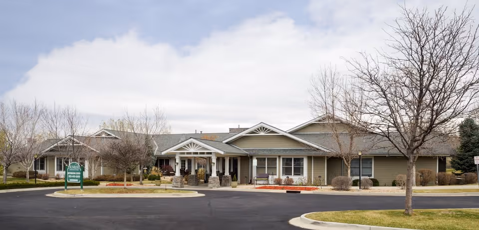 Exterior view of a single-story senior living facility building with a covered entrance, leafless trees, and a circular driveway. The sky is partly cloudy.