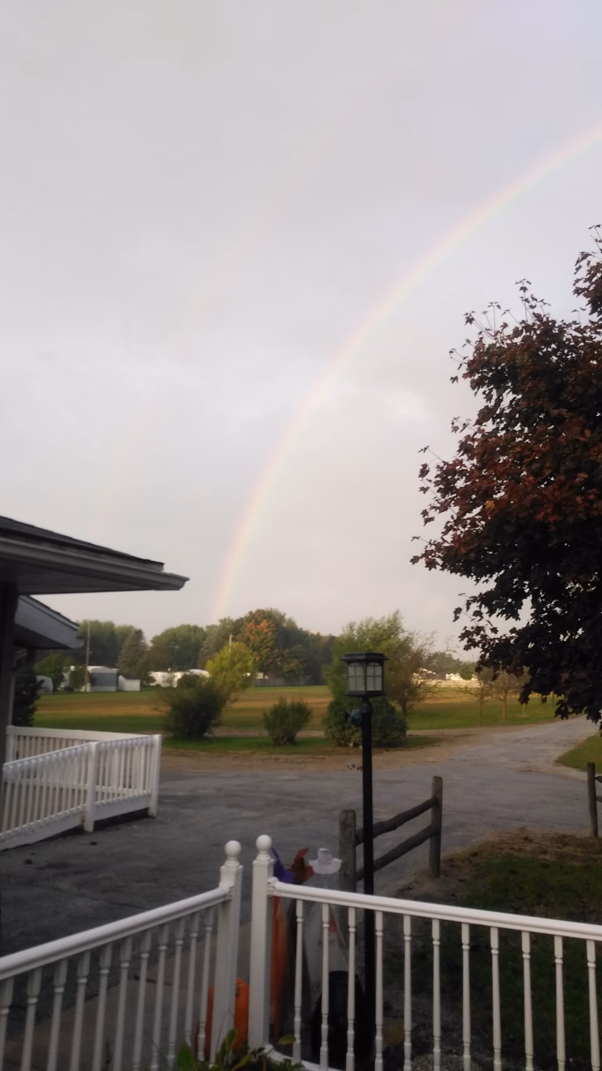 View from a porch or balcony overlooking a paved driveway and grassy area with trees. A rainbow is visible in the sky above the landscape. There is a white railing in the foreground and a black lamp post near the center of the image.