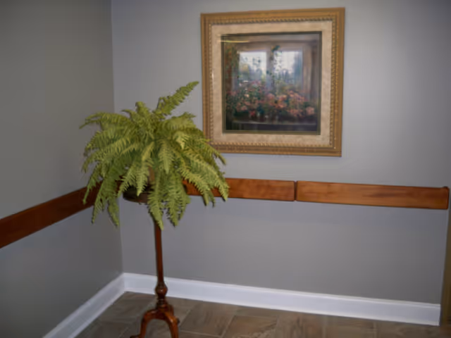 Corner of a room with gray walls, a wooden handrail, a framed painting of flowers, and a tall potted fern on a wooden stand.