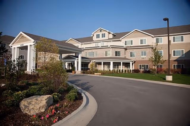 Exterior view of a senior living facility named Senior Star at Elmore Place, showing a large multi-story building with a covered entrance, landscaped garden with bushes and flowers, and a driveway under a clear blue sky.