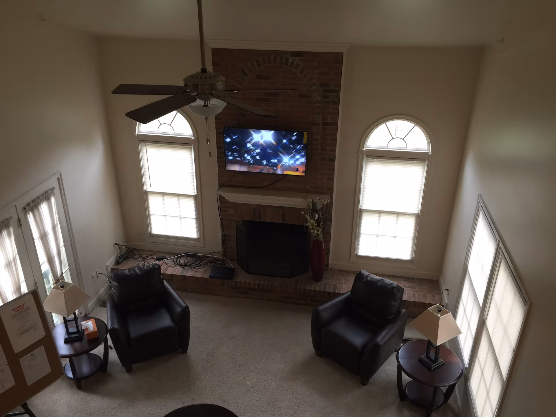 View from above of a living room with two black armchairs facing a brick fireplace with a mounted flat-screen TV above it. The room has two tall arched windows with blinds on either side of the fireplace, a ceiling fan with lights, two side tables with lamps, and a bulletin board near a set of French doors with sheer curtains.