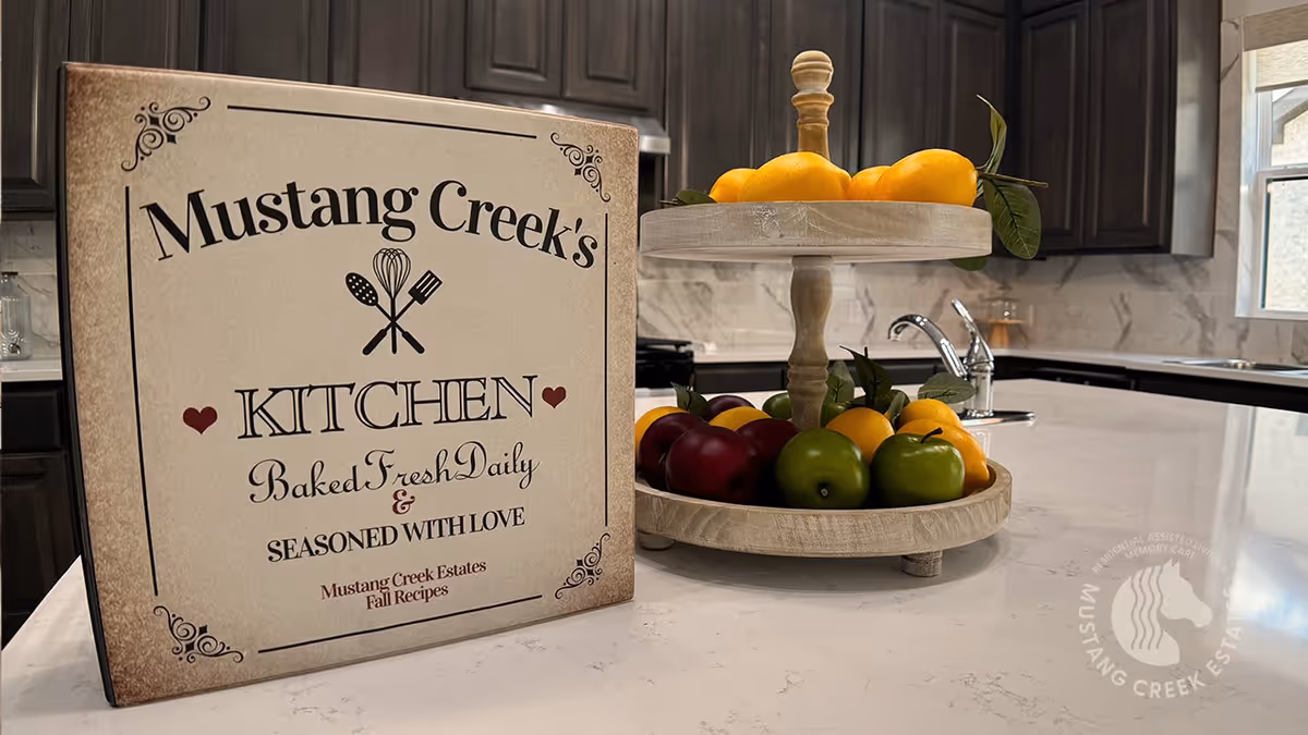 A kitchen countertop with a decorative sign that reads 'Mustang Creek's Kitchen Baked Fresh Daily & Seasoned With Love Mustang Creek Estates Fall Recipes' next to a two-tiered wooden fruit stand holding lemons, apples, and other fruits. The background shows dark wooden cabinets and a window.