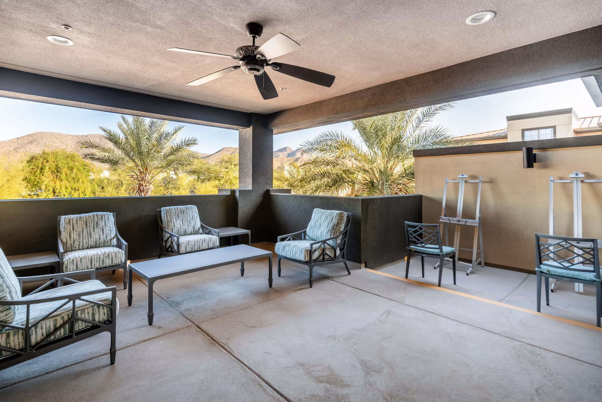 Covered outdoor patio area with cushioned chairs, a coffee table, ceiling fan, and easels. The patio overlooks palm trees and distant mountains under a clear sky.