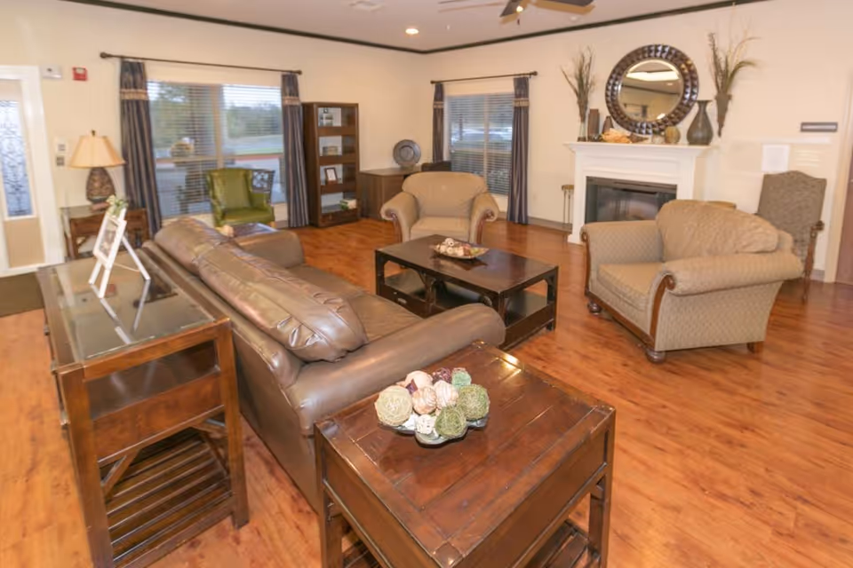 Communal living room with a leather sofa, armchairs, coffee table, and a fireplace in a senior facility.