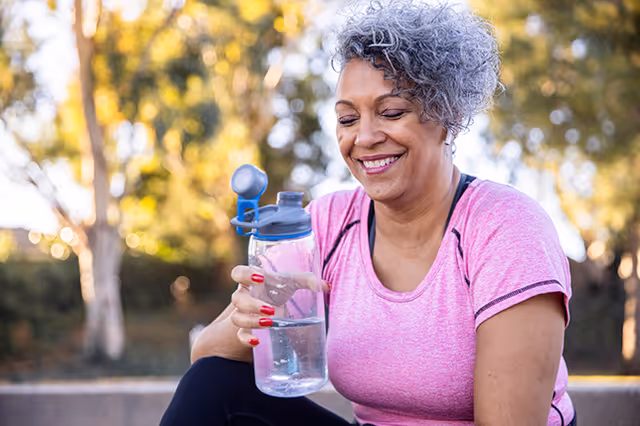 A smiling older woman with curly gray hair wearing a pink athletic shirt is holding a clear water bottle with a blue lid while sitting outdoors with trees and sunlight in the background.