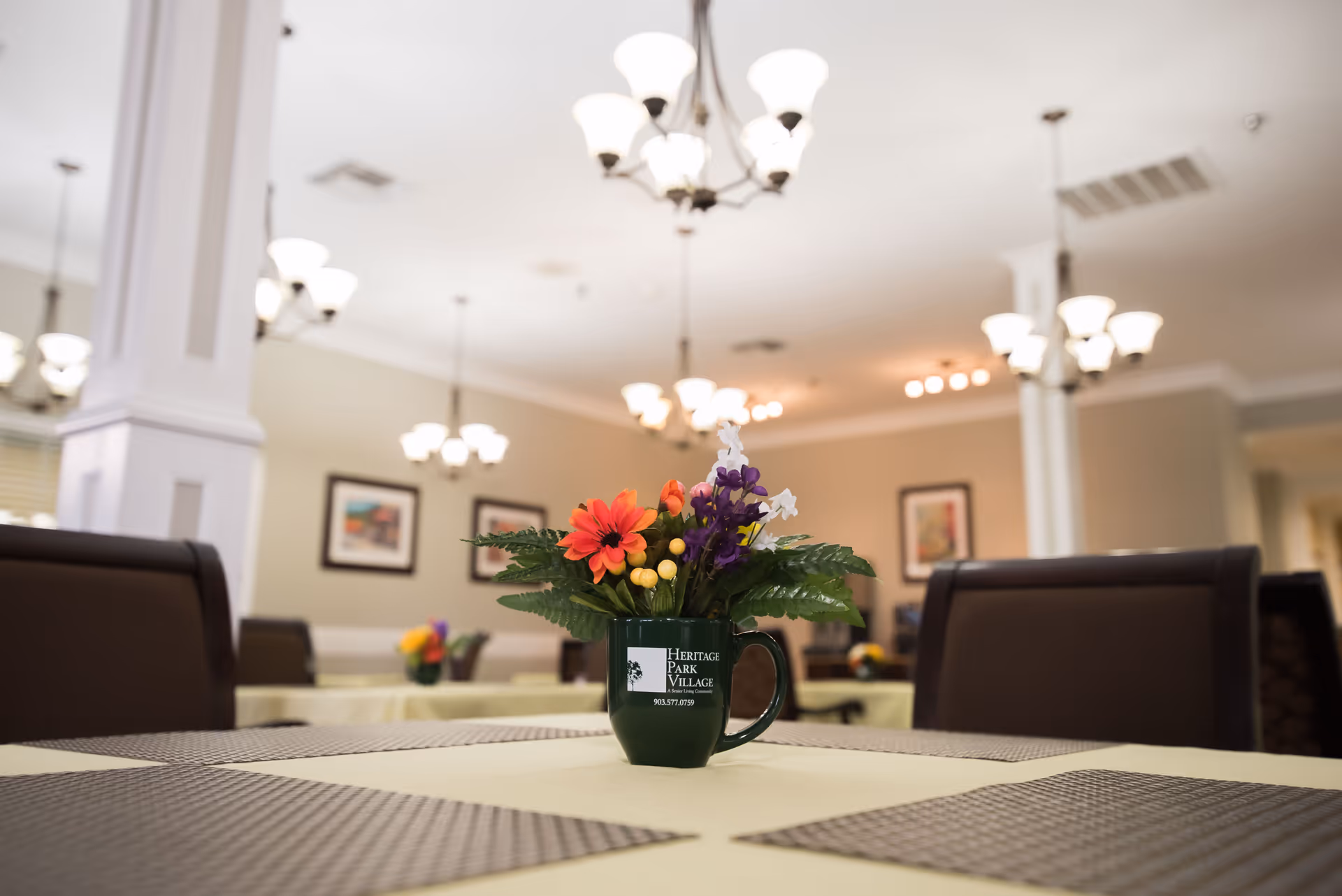 Interior view of a dining room in an assisted living facility with a table centerpiece featuring a green mug filled with colorful artificial flowers. The mug has the Heritage Park Village logo and contact number. The room has multiple tables with placemats, brown chairs, framed artwork on the walls, and several ceiling light fixtures.