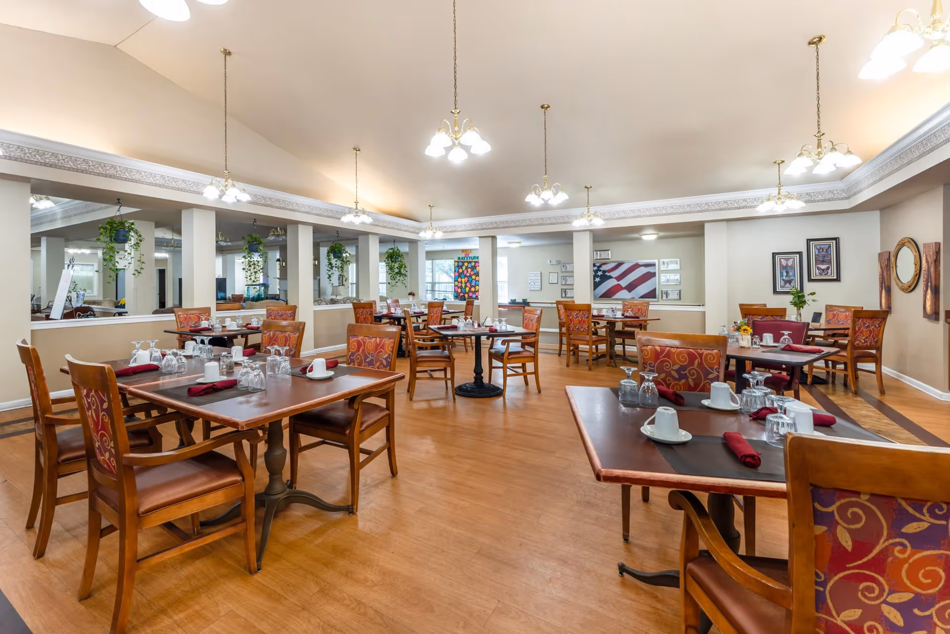 Spacious communal dining room with multiple set wooden tables and chairs, hanging plants, and chandelier lighting.