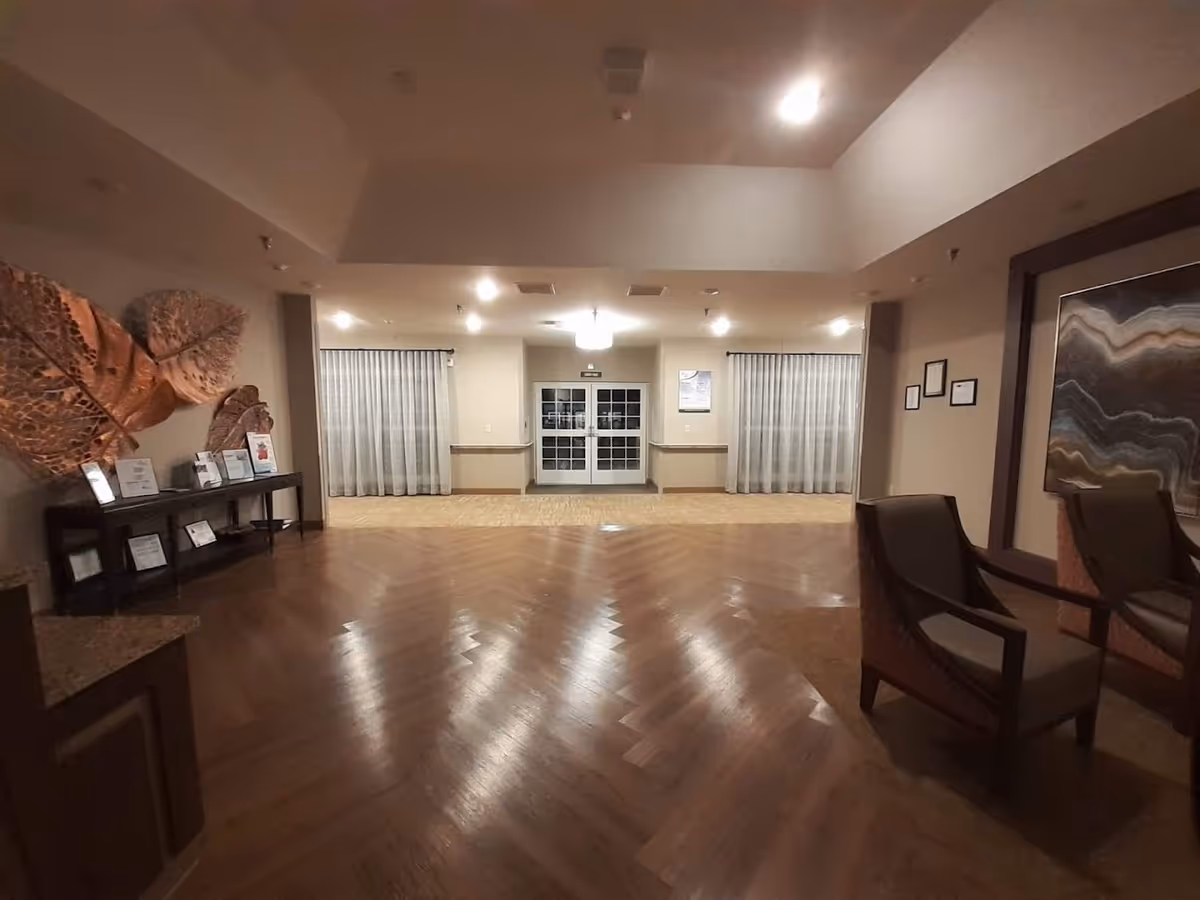 Interior view of a nursing and rehabilitation center lobby area with wooden flooring, two armchairs on the right, a large decorative wall art, a console table with framed certificates and decorations on the left, and double glass doors at the far end flanked by windows with sheer curtains.