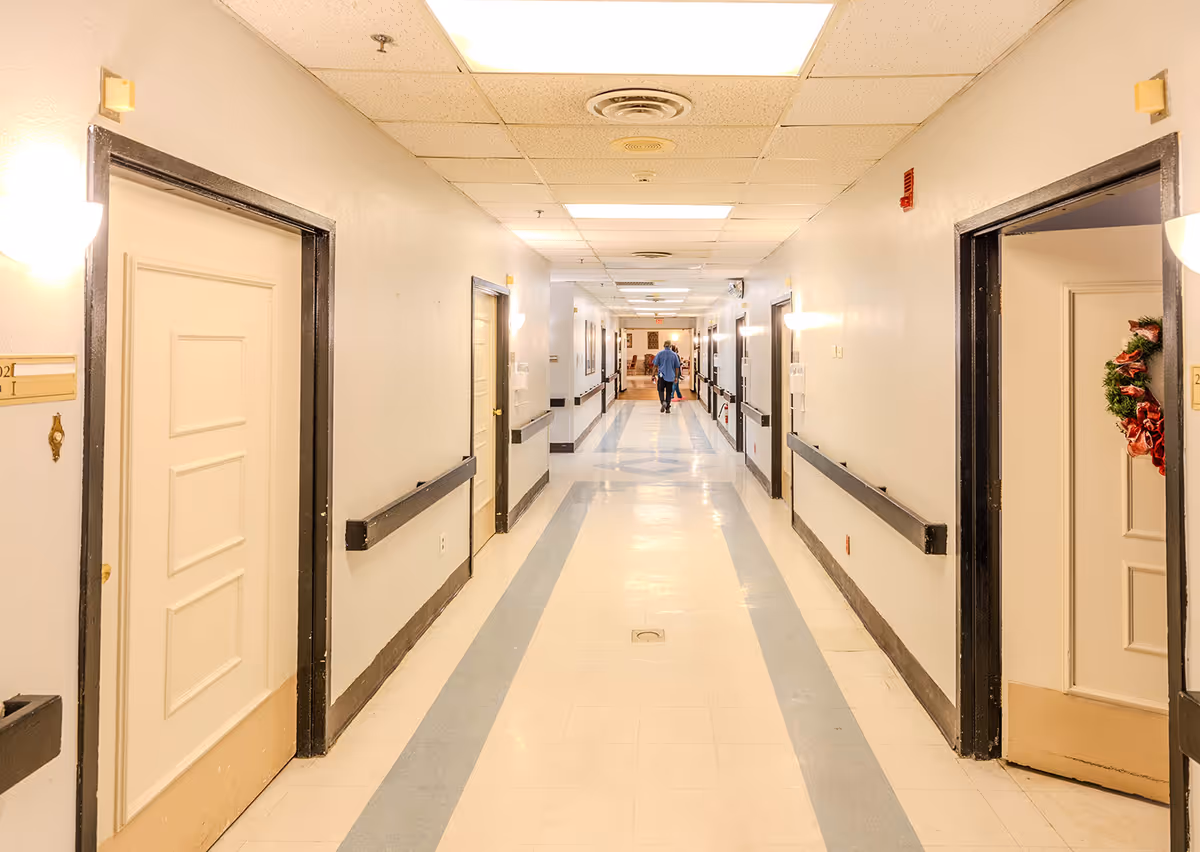 Long, well-lit interior hallway in a senior living facility with doors on both sides and a person walking away in the distance.