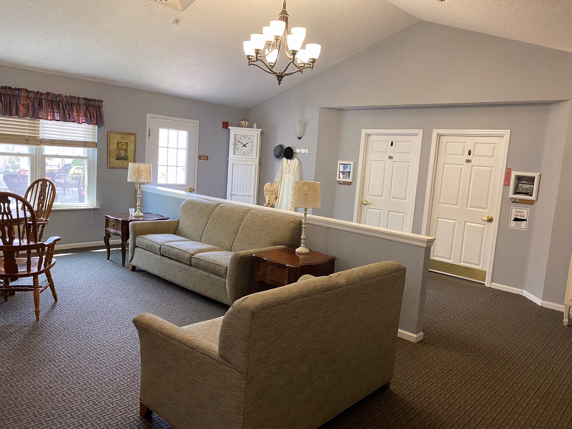 Seating area in a senior living common room with sofas, side tables and lamps, a dining table by a window, and hallway doors.