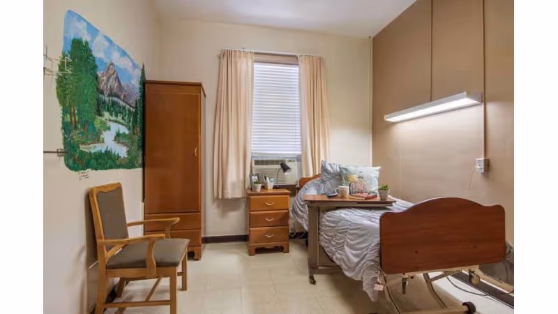 A small, simple nursing facility bedroom with a single hospital-style bed, a wooden chair, a wooden wardrobe, a small wooden nightstand with a lamp and some items on it, and a window with beige curtains and a window air conditioning unit. There is a colorful mural of a nature scene with trees, mountains, and a river painted on one wall.