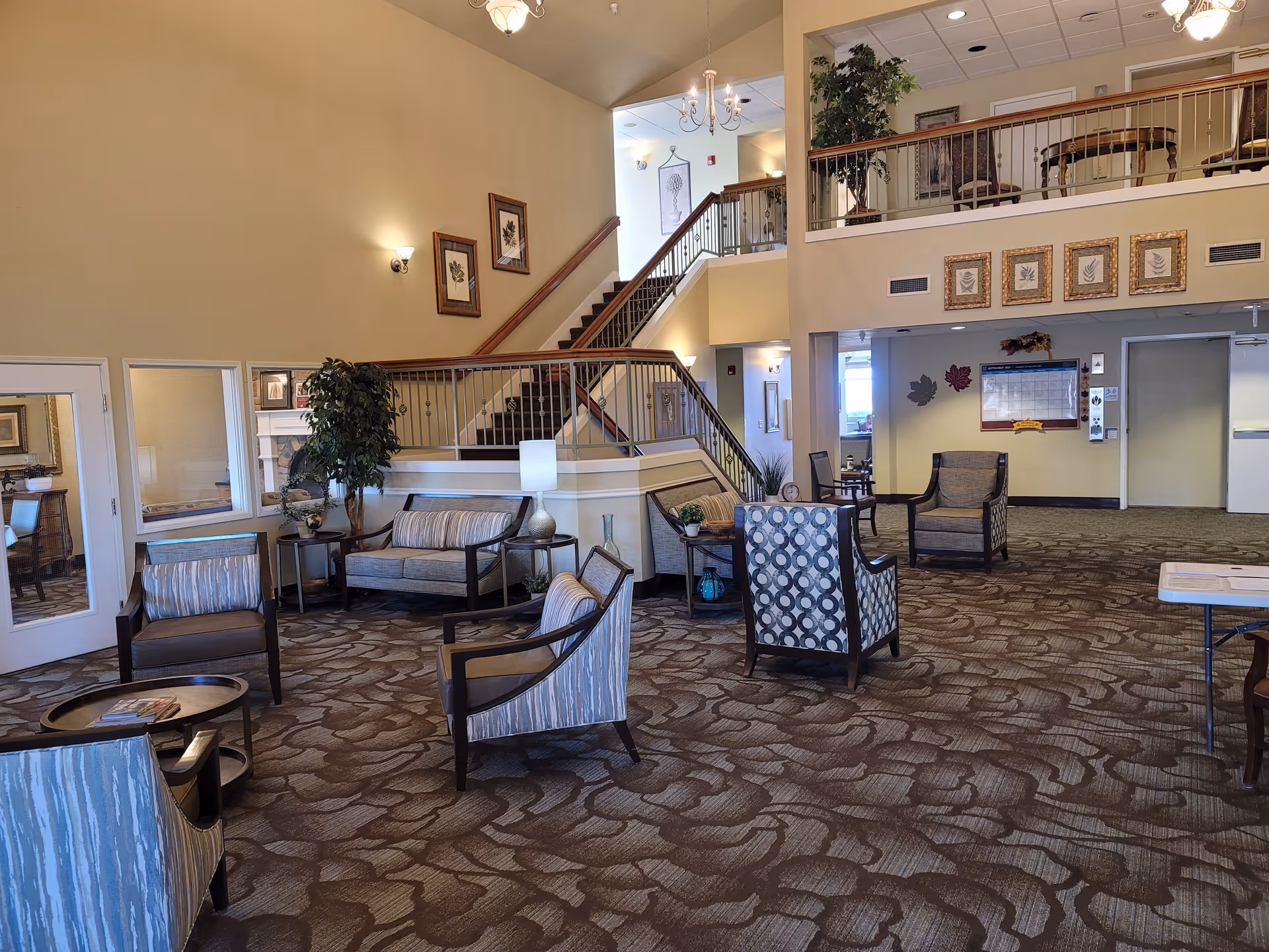 A spacious senior living facility common area with patterned carpet, multiple armchairs and small tables arranged for seating. There is a staircase leading to an upper level with a railing and additional seating. The walls are decorated with framed artwork and there are plants and lamps placed around the room. The area is well-lit with wall sconces and ceiling lights.