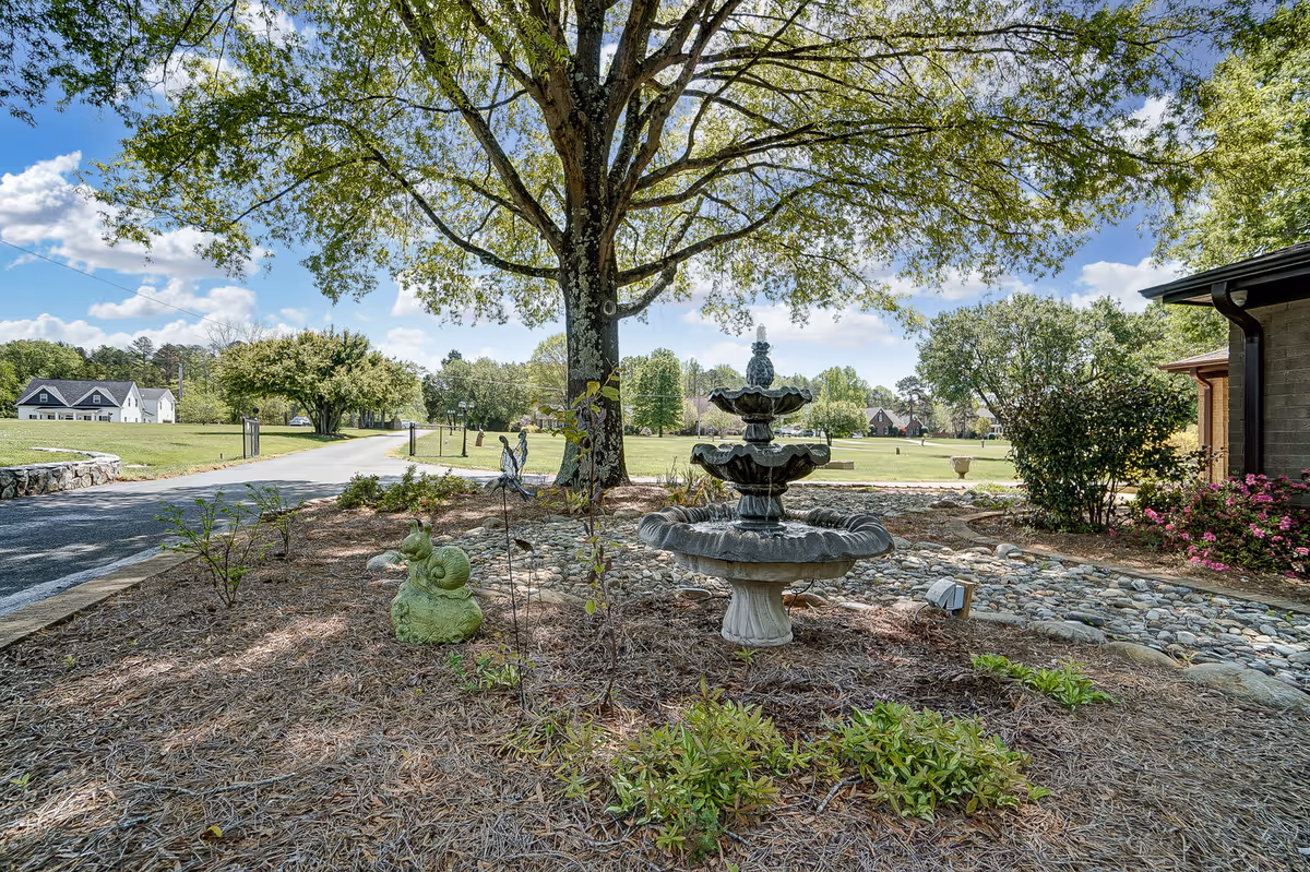 Three-tier stone fountain in a landscaped garden bed under a large tree with open lawns and houses in the background.