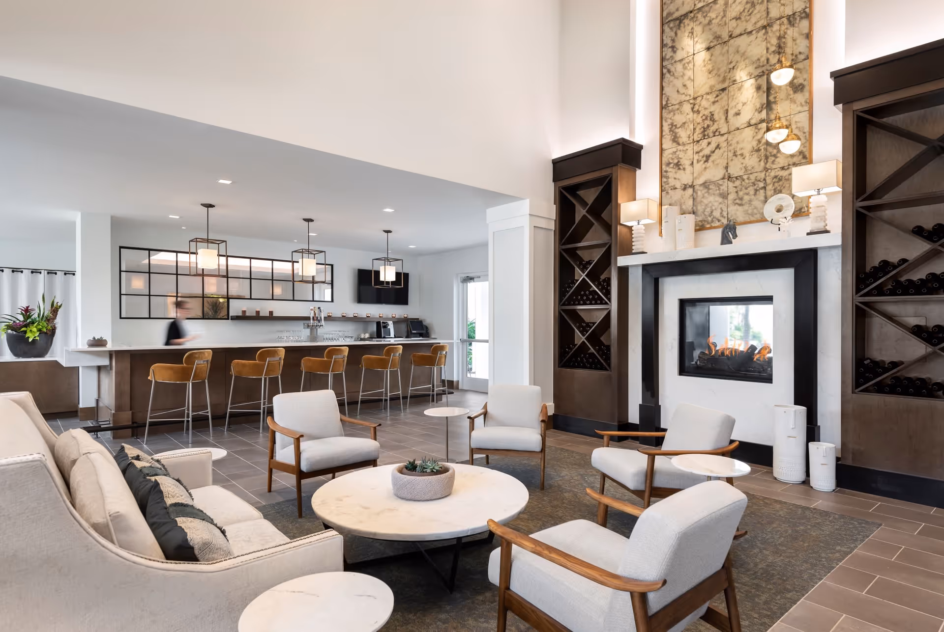 A modern and cozy common area with a fireplace surrounded by built-in wine racks, a large decorative mirror above the fireplace, and comfortable seating including a sofa and four armchairs around a round coffee table. In the background, there is a bar counter with high stools and pendant lights hanging from the ceiling.