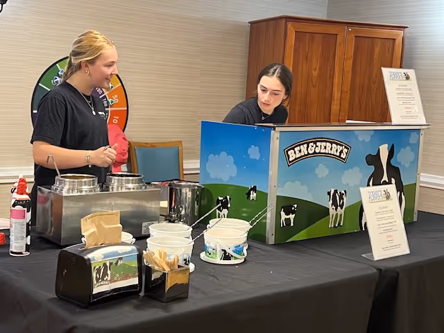 Two women standing behind a table with a Ben & Jerry's ice cream cart and various ice cream serving supplies in a room with beige walls and a wooden cabinet in the background.