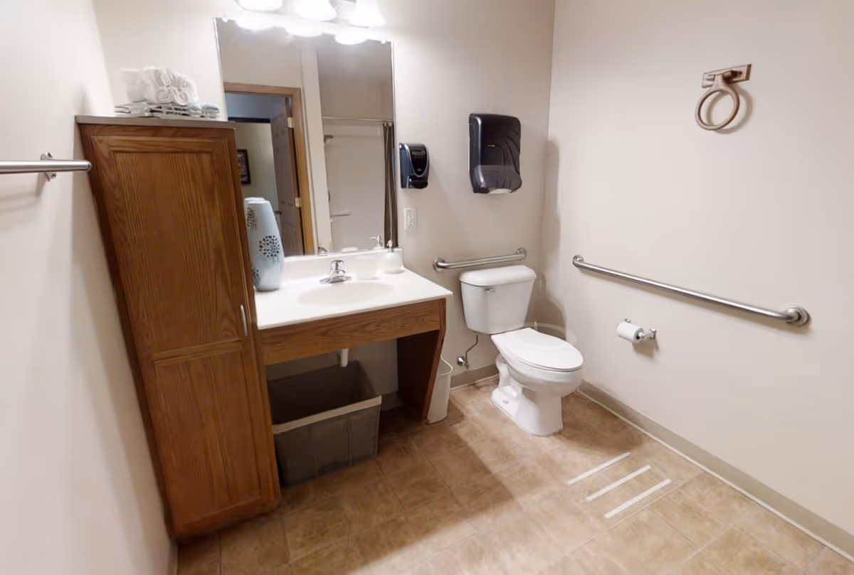 A clean and accessible bathroom with a wooden cabinet, a sink with a large mirror above it, a toilet with grab bars on the walls, a paper towel dispenser, and a trash bin underneath the sink. The floor is tiled and the walls are painted white.