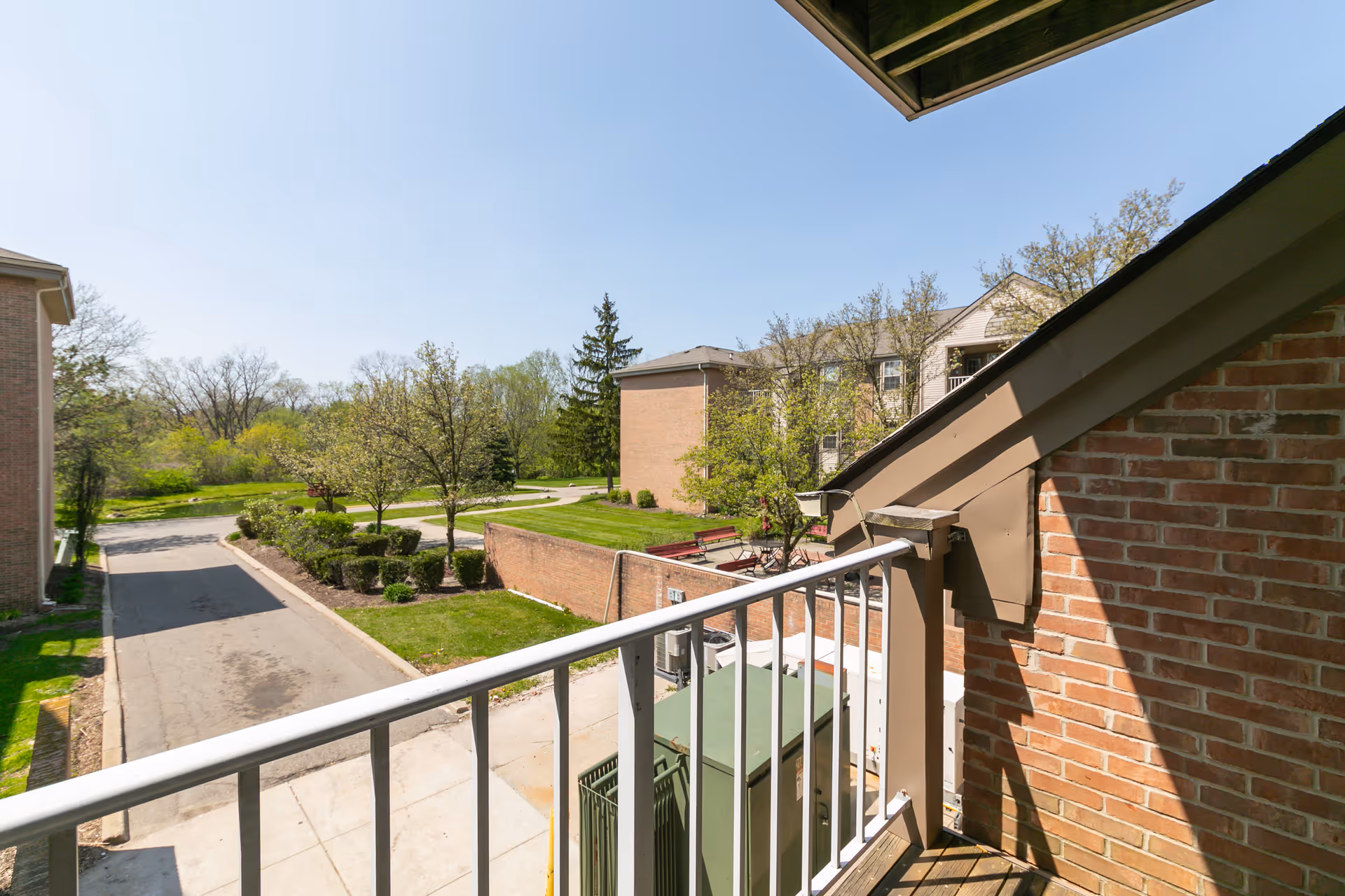 View from a second-floor balcony over a driveway and landscaped courtyard with brick buildings and trees under a clear blue sky.
