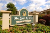 Outdoor view of a sign for Life Care Center of Haltom surrounded by green bushes and flowers with a partly cloudy sky in the background.