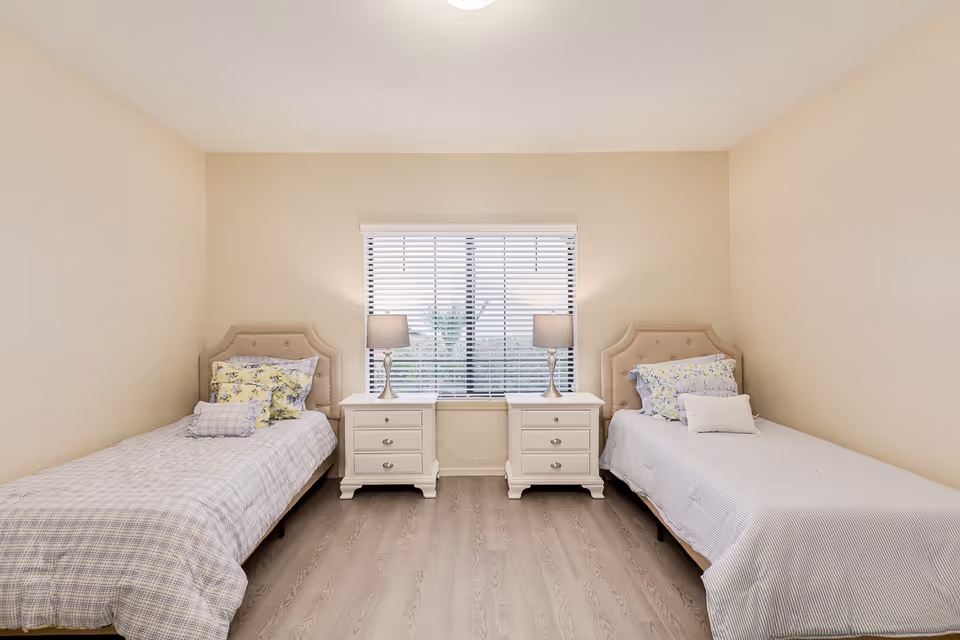 A simple bedroom with two single beds placed on either side of a window. Each bed has a beige upholstered headboard and is made with light-colored bedding and decorative pillows. Between the beds are two white nightstands, each with a lamp. The room has light-colored walls and wood flooring.
