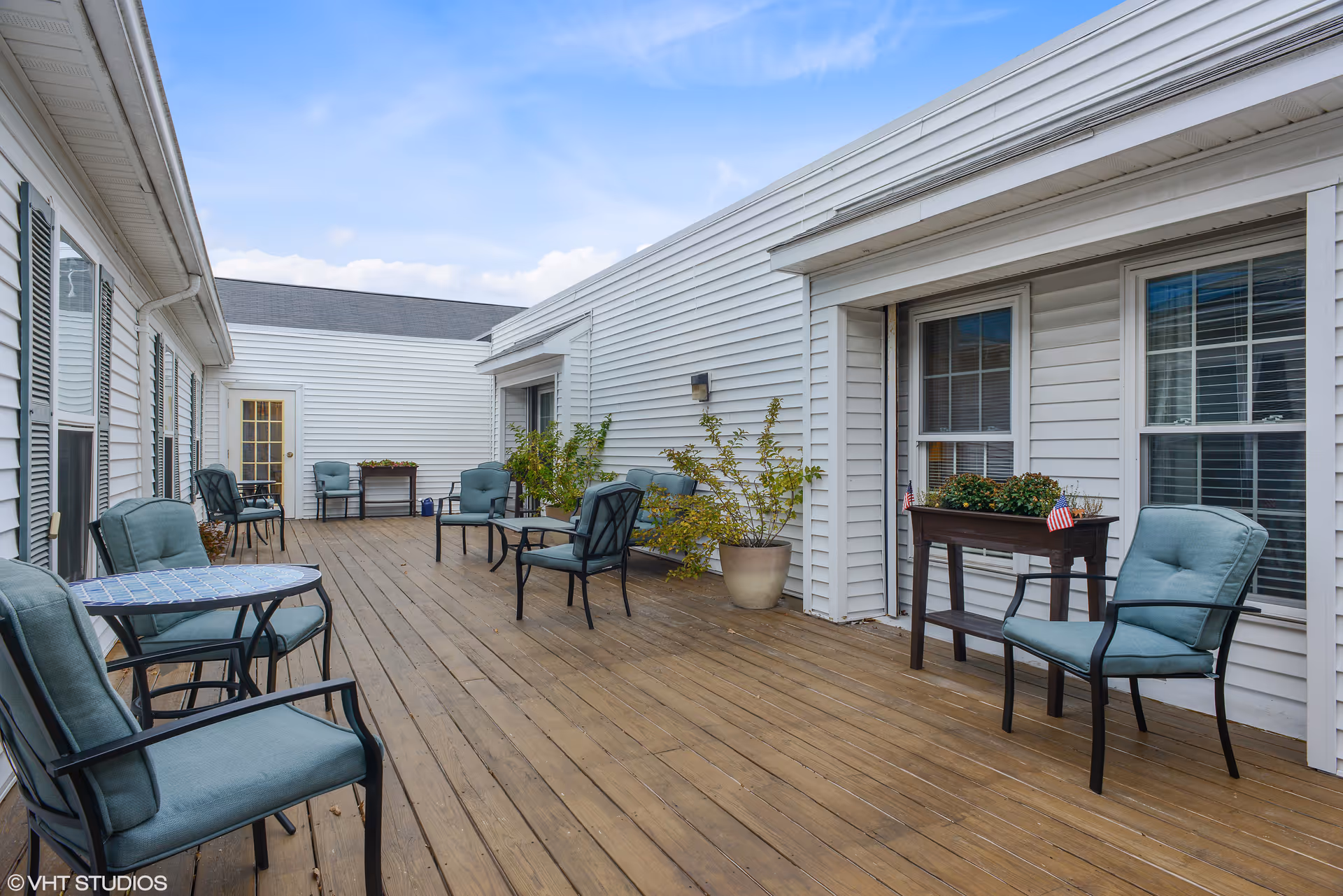 Outdoor wooden deck patio with cushioned chairs, small tables, potted plants and white siding.