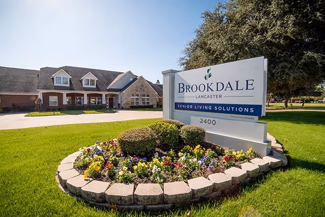 Exterior view of Brookdale Lancaster senior living facility with a large sign in the foreground surrounded by a flower bed and green lawn, and the building visible in the background under a clear blue sky.