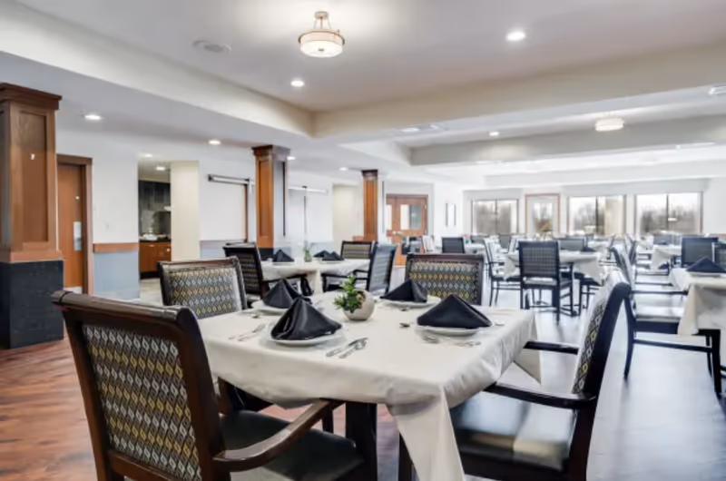 A spacious dining room in Arbor Glen Senior Living with multiple tables covered in white tablecloths, each set with black folded napkins, plates, and silverware. The room features wooden chairs with patterned upholstery, large windows letting in natural light, and wooden columns with a modern ceiling light fixture.