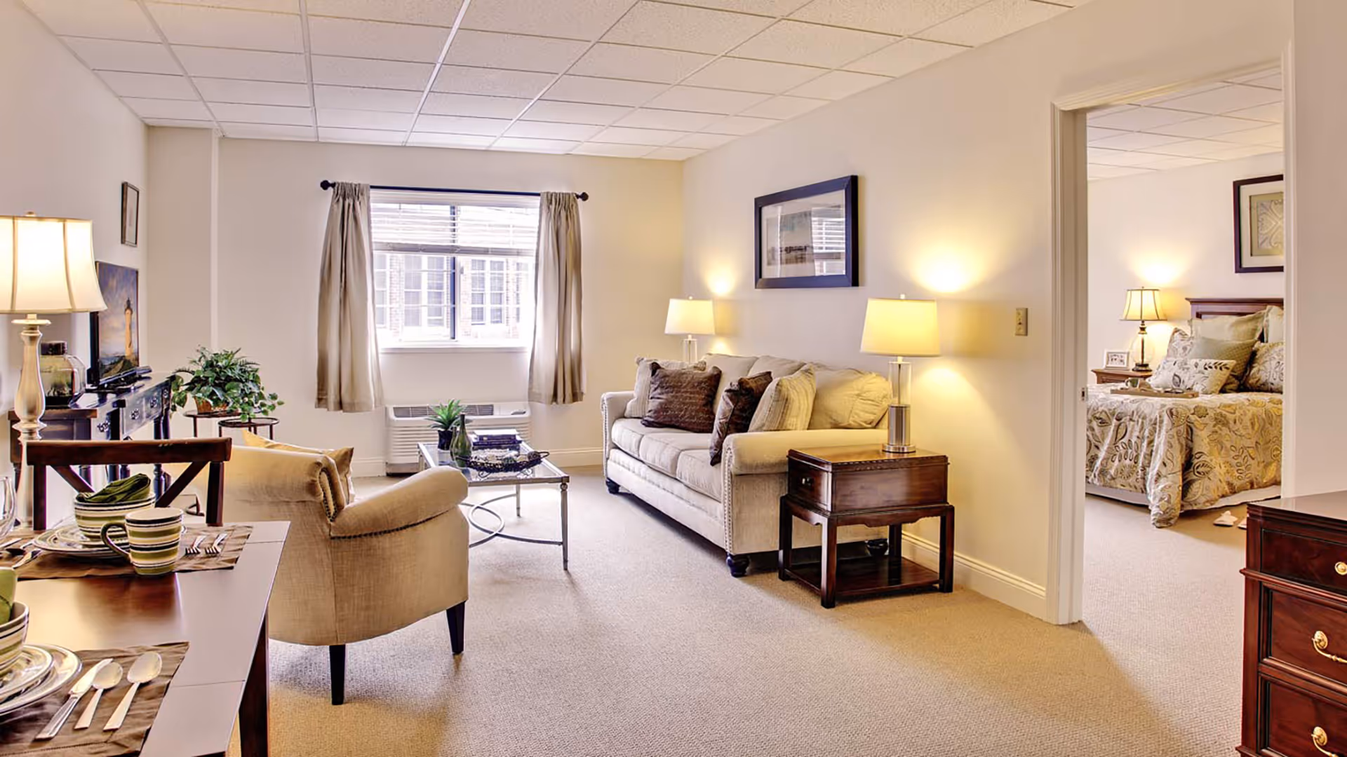 Well-lit living room with a sofa, armchair, coffee table and dining area, with a bedroom visible through an open doorway.