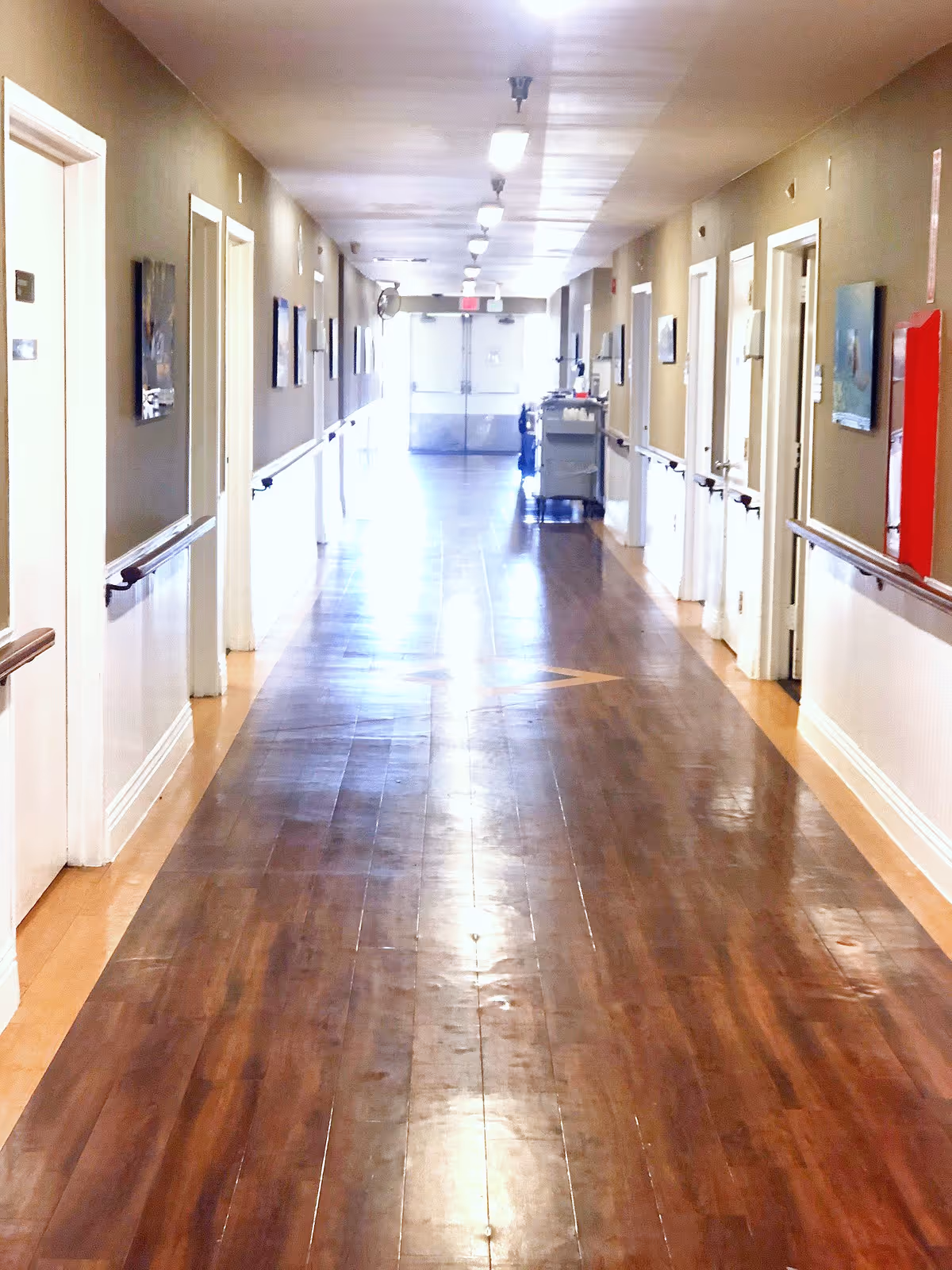 A long, well-lit hallway in a care facility with wooden flooring and beige walls. There are multiple doors on both sides of the hallway, handrails along the walls, and some framed pictures hanging. At the far end, double doors with exit signs are visible, along with a cleaning cart on the right side.