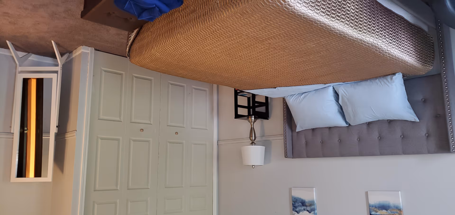 A bedroom with a bed featuring a brown textured bedspread and two light blue pillows. The bed has a tufted gray headboard with nailhead trim. Next to the bed is a silver lamp with a white lampshade on a small black nightstand. There is a white closet with double doors and a white vanity mirror with a small stool in the corner. Two framed abstract paintings hang on the wall above the bed.