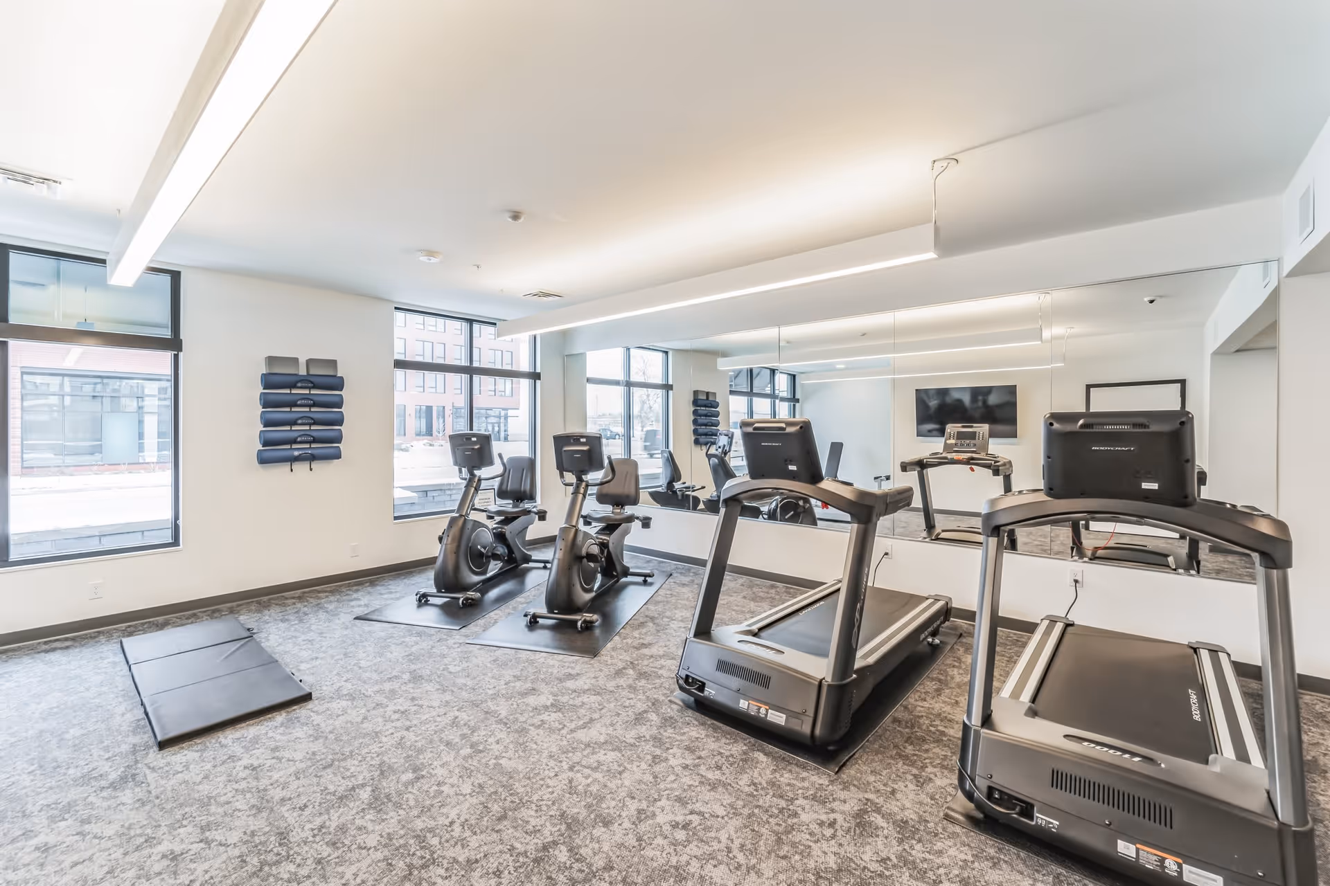 Bright fitness room with treadmills, stationary bikes, exercise mats, and a large mirror on the wall.