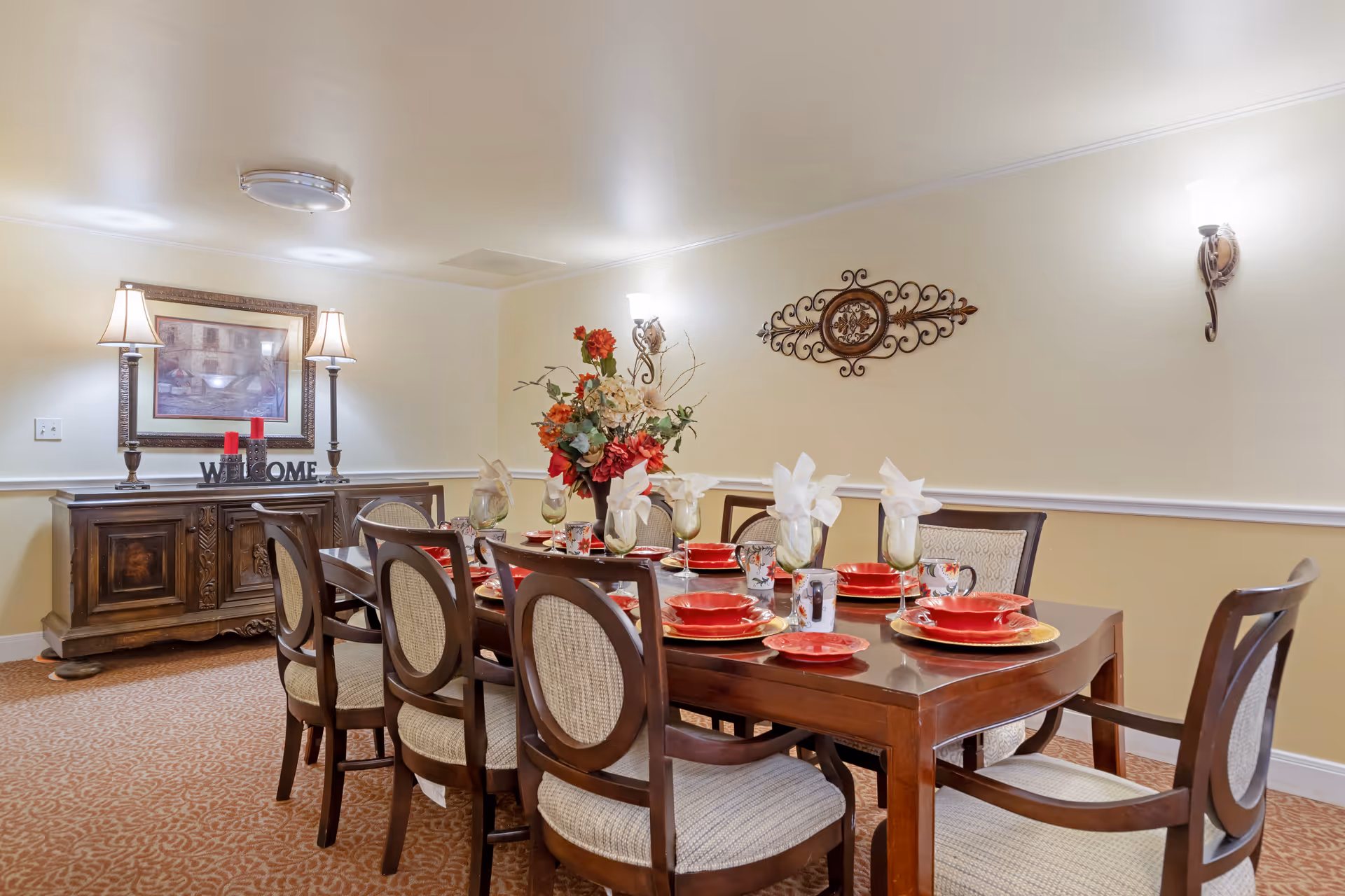 A dining room with a long wooden table set for eight people. The table is decorated with red plates, cups, and folded white napkins in glasses. A floral centerpiece with red and white flowers is in the middle of the table. The room has beige walls with two wall sconces and a framed picture above a wooden sideboard that holds two lamps and a decorative 'WELCOME' sign with red candles.