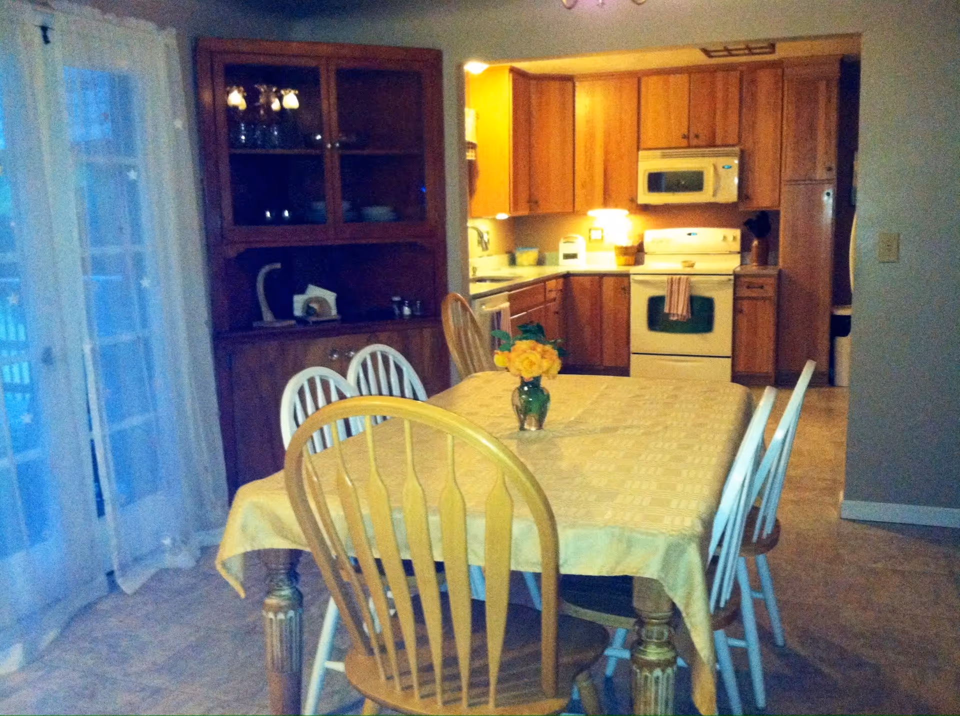A dining area with a wooden table covered by a yellow tablecloth and six chairs around it, some wooden and some white. A small vase with yellow flowers is placed in the center of the table. Behind the dining area is a kitchen with wooden cabinets, a white stove with a towel hanging on the handle, a microwave above the stove, and a refrigerator to the right. To the left of the dining area is a wooden cabinet with glass doors and a sheer curtain covering a sliding glass door.