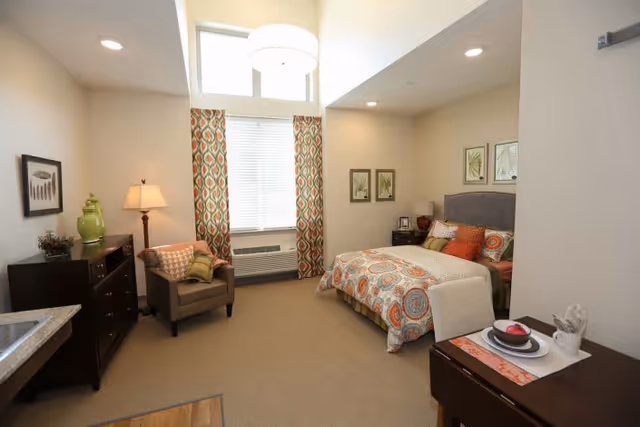 A cozy bedroom in a senior living facility with a bed featuring colorful patterned bedding and multiple pillows. The room includes a cushioned armchair with a pillow, a wooden dresser with decorative items, a floor lamp, and framed botanical artwork on the walls. There is a large window with patterned curtains letting in natural light, and a small table set with a plate, bowl, and napkin. The room has a warm, inviting atmosphere.
