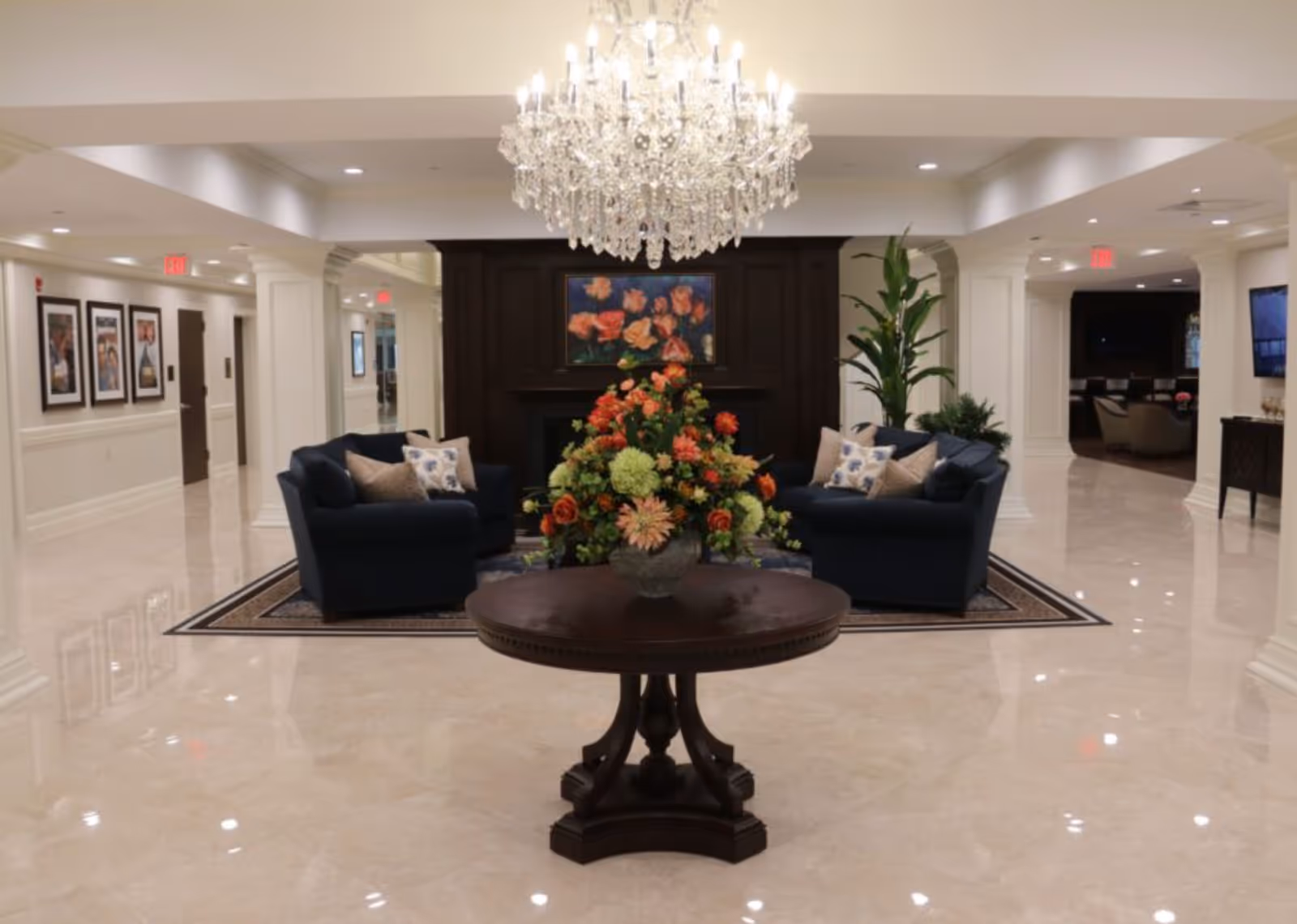 A spacious and elegant common area with polished marble floors, a large crystal chandelier hanging from the ceiling, a round wooden table with a floral arrangement in the center, two navy blue sofas with decorative pillows arranged around a dark wood fireplace, and framed artwork on the walls.