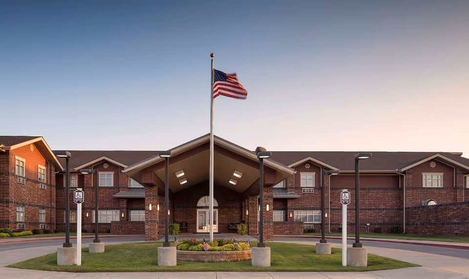 Front exterior view of Canoe Brook Assisted Living - Duncan building with a flagpole flying the American flag in the center of a circular driveway, surrounded by well-maintained landscaping and handicap parking signs.