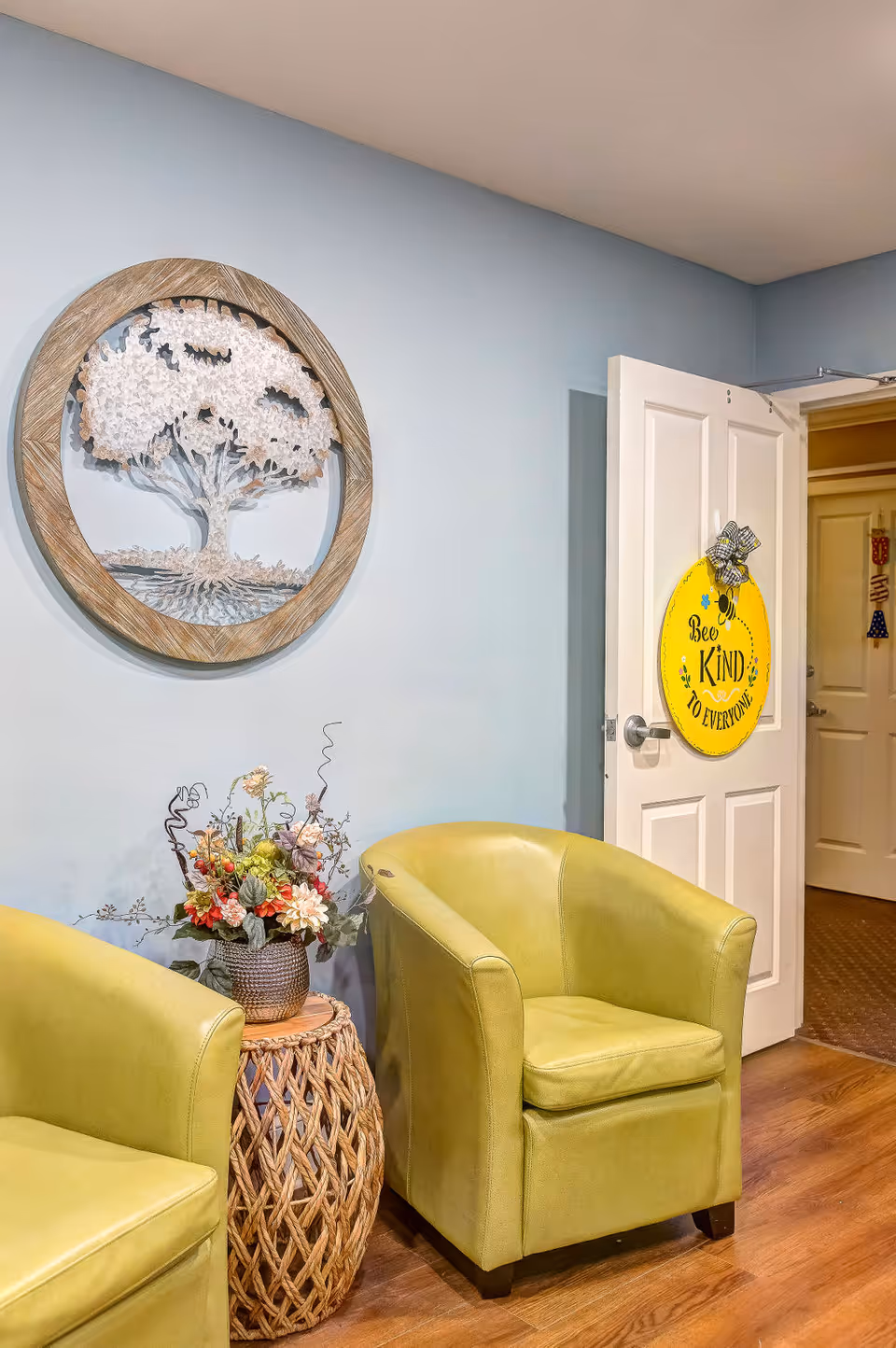 A cozy seating area with two light green armchairs and a woven side table holding a floral arrangement. On the light blue wall above the chairs is a round wooden-framed artwork of a tree. A white door is open, displaying a yellow circular sign that reads 'Bee Kind to Everyone'.