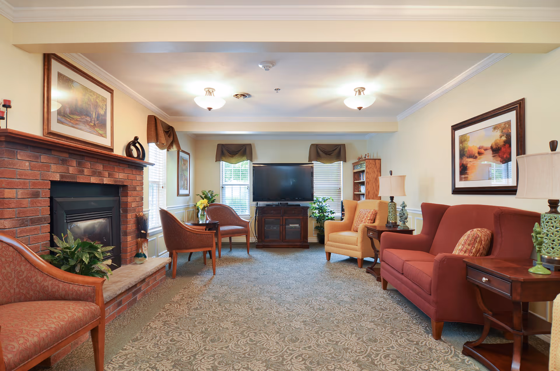 A cozy common living room with armchairs and sofas arranged around a brick fireplace and a television.