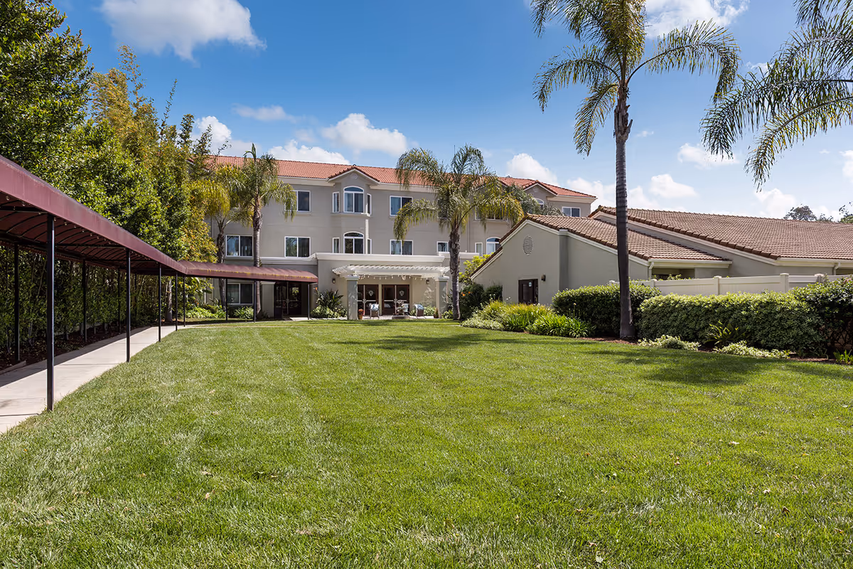 A well-maintained outdoor lawn area with palm trees and bushes, adjacent to a multi-story building with a red-tiled roof and beige exterior. There is a covered walkway on the left side and a patio area with seating near the building entrance under a pergola.