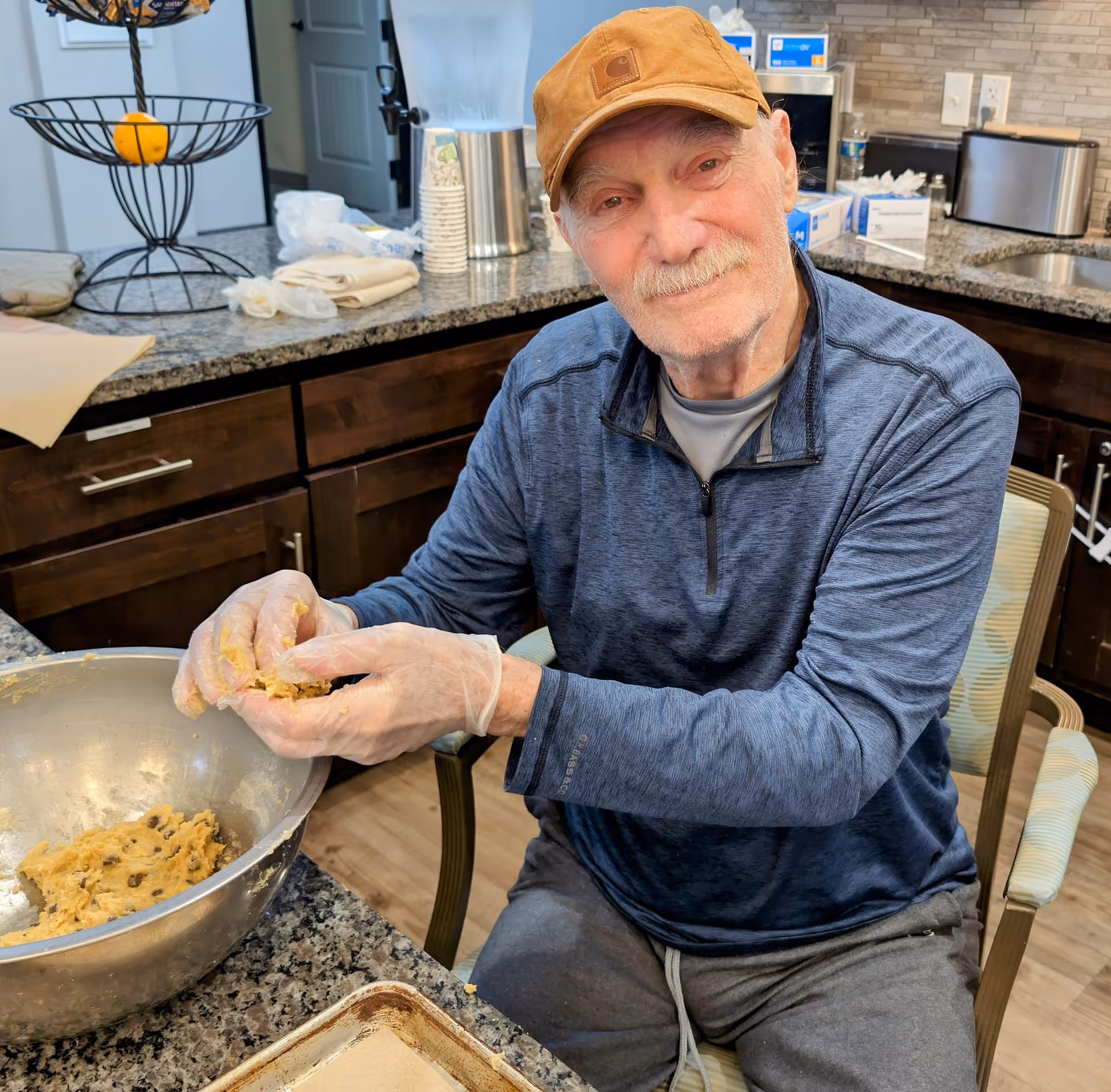 An elderly man wearing a brown cap and blue long-sleeve shirt is sitting at a kitchen counter, shaping cookie dough with gloved hands. There is a large metal mixing bowl with cookie dough and a baking tray on the granite countertop. The background shows kitchen cabinets, a sink, and various kitchen items.
