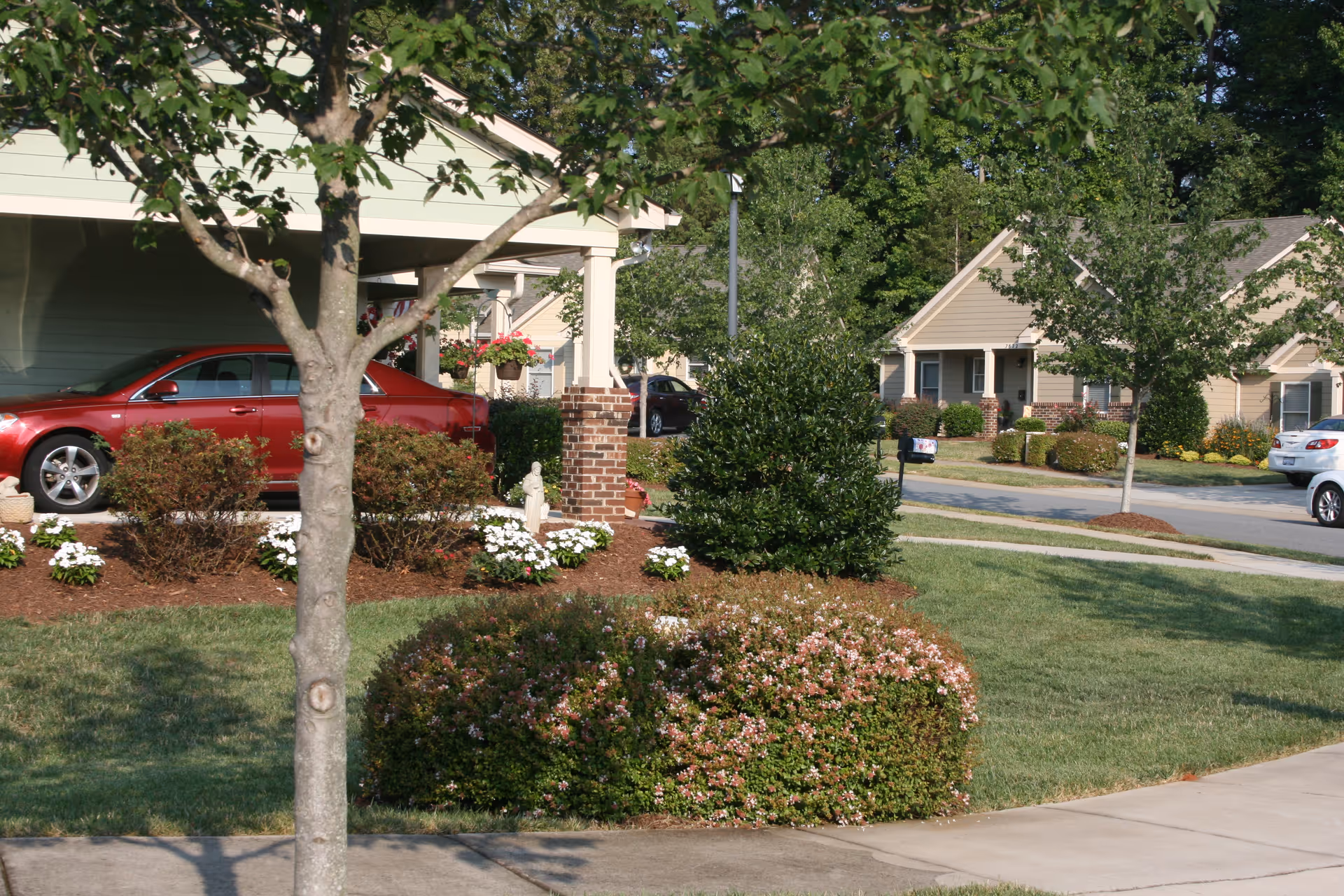 View of a residential neighborhood with well-maintained lawns, bushes, and trees. Several single-story houses with porches and parked cars are visible along a paved street under a clear sky.