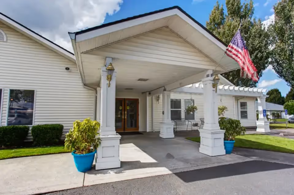 Front entrance of a white building with a covered driveway supported by white columns. There are two blue pots with green plants on either side of the entrance. An American flag is mounted on the right side of the entrance. The building is surrounded by green grass and trees under a partly cloudy sky.