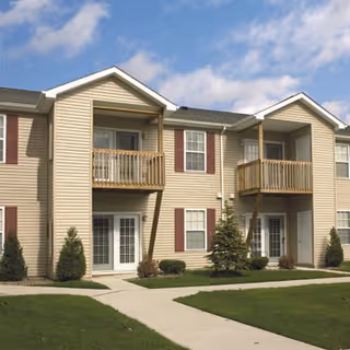 Exterior view of a two-story senior apartment building with beige siding, brown shutters, and small balconies. There is a well-maintained lawn and a concrete walkway leading to the entrances under a partly cloudy sky.