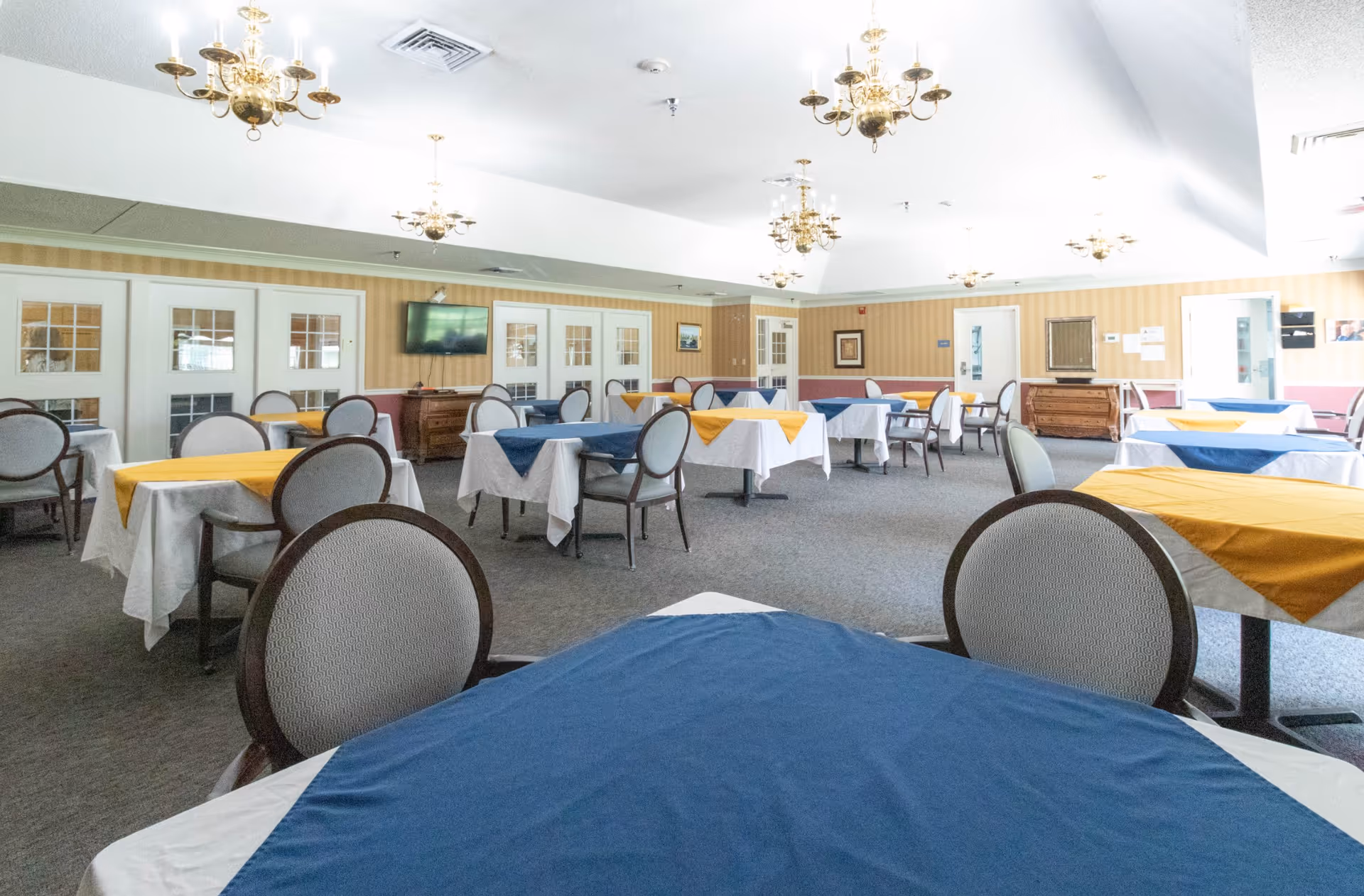 A spacious dining room with multiple tables covered in white tablecloths and blue or yellow table runners. Each table is surrounded by cushioned chairs with rounded backs. The room has beige striped wallpaper with a maroon lower border, several brass chandeliers hanging from the ceiling, and a flat-screen TV mounted on the far wall above a wooden cabinet. There are multiple white doors with glass panes along the walls.