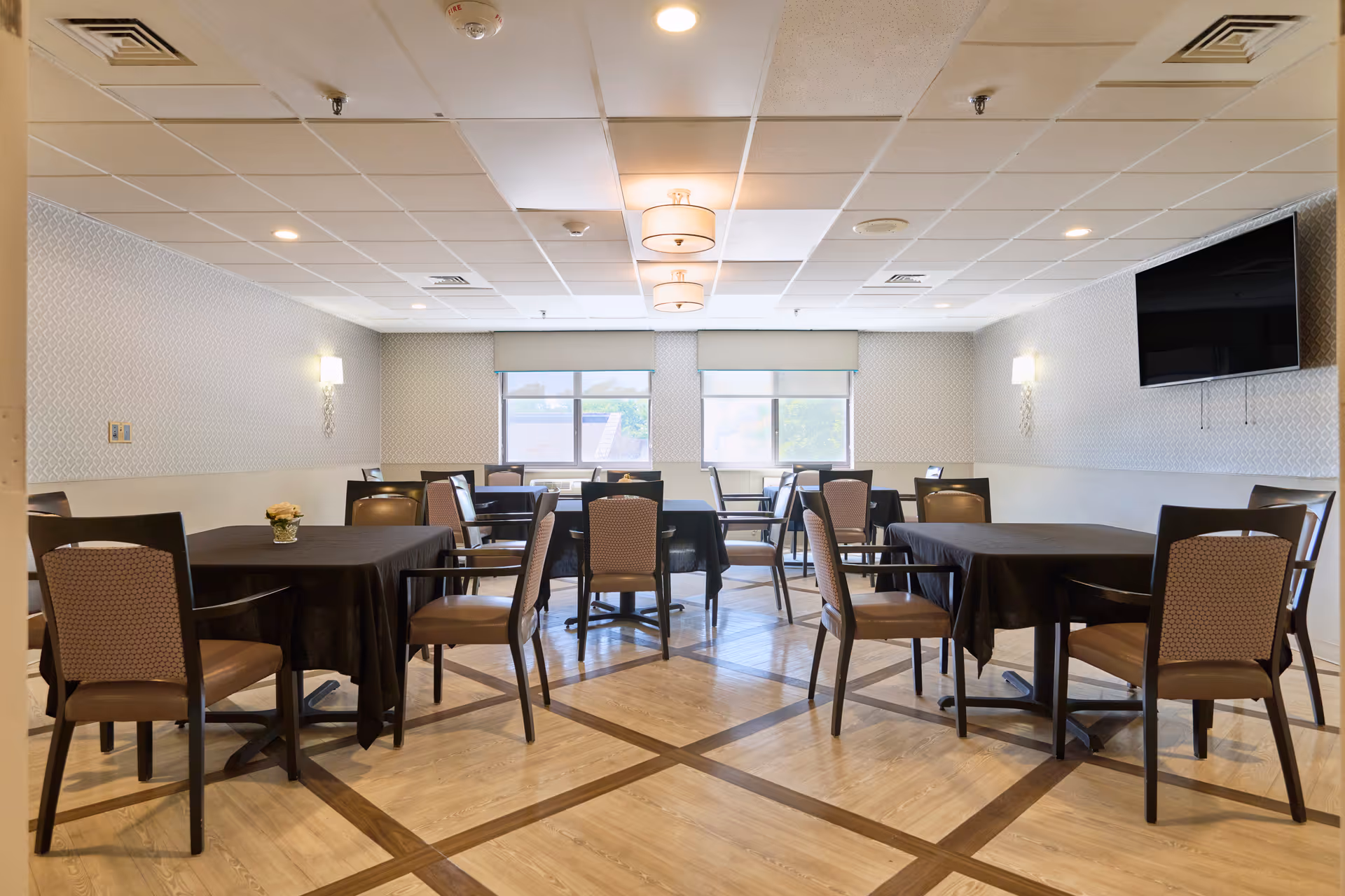 A well-lit dining room with several tables covered in black tablecloths and surrounded by brown cushioned chairs. The room has light-colored walls with patterned wallpaper, two windows with blinds, ceiling lights, wall sconces, and a flat-screen TV mounted on the right wall. The floor has a wood pattern with darker wood inlays.
