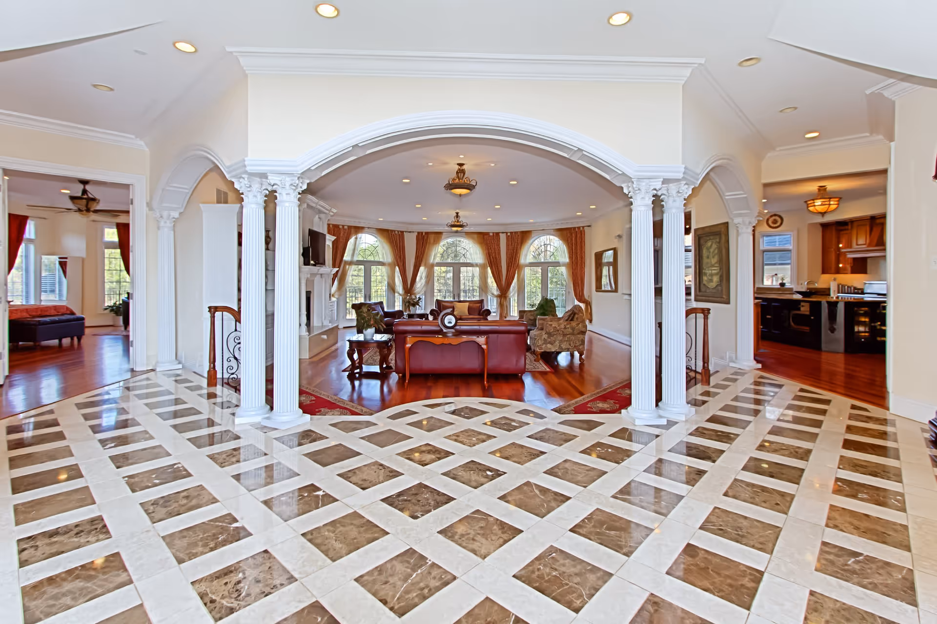 Spacious and elegant interior of a senior living facility featuring a polished marble floor with a checkered pattern, white decorative columns and arches, a living room area with leather and upholstered seating, large windows with orange curtains, and adjacent rooms including a bedroom and a kitchen.