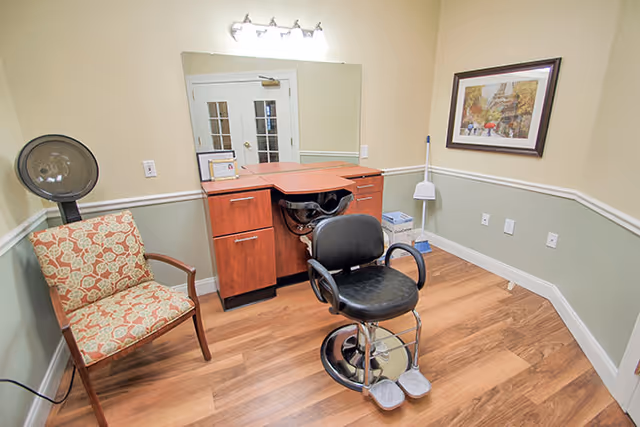 Interior view of a salon area in a senior living facility with a black salon chair in front of a wooden cabinet with a built-in sink, a patterned armchair to the left, a large mirror on the wall, a framed picture on the right wall, and a broom and dustpan in the corner.